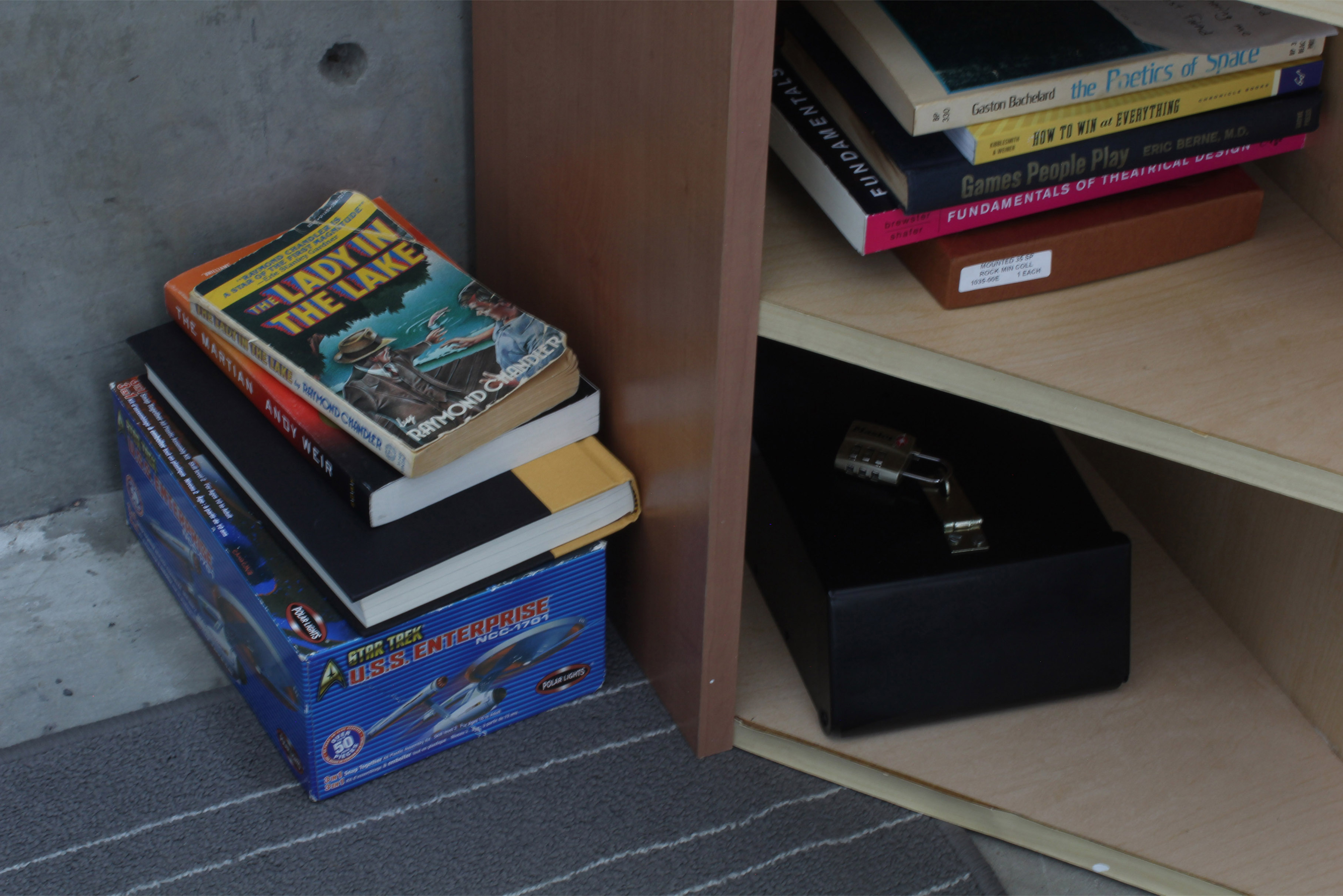 Bottom-half of a bookshelf. Next to it on the floor is a stack of books and a box of a model Star Trek spaceship. On the shelf are books about space, games and theatrical design. On the bottom shelf is a lockbox