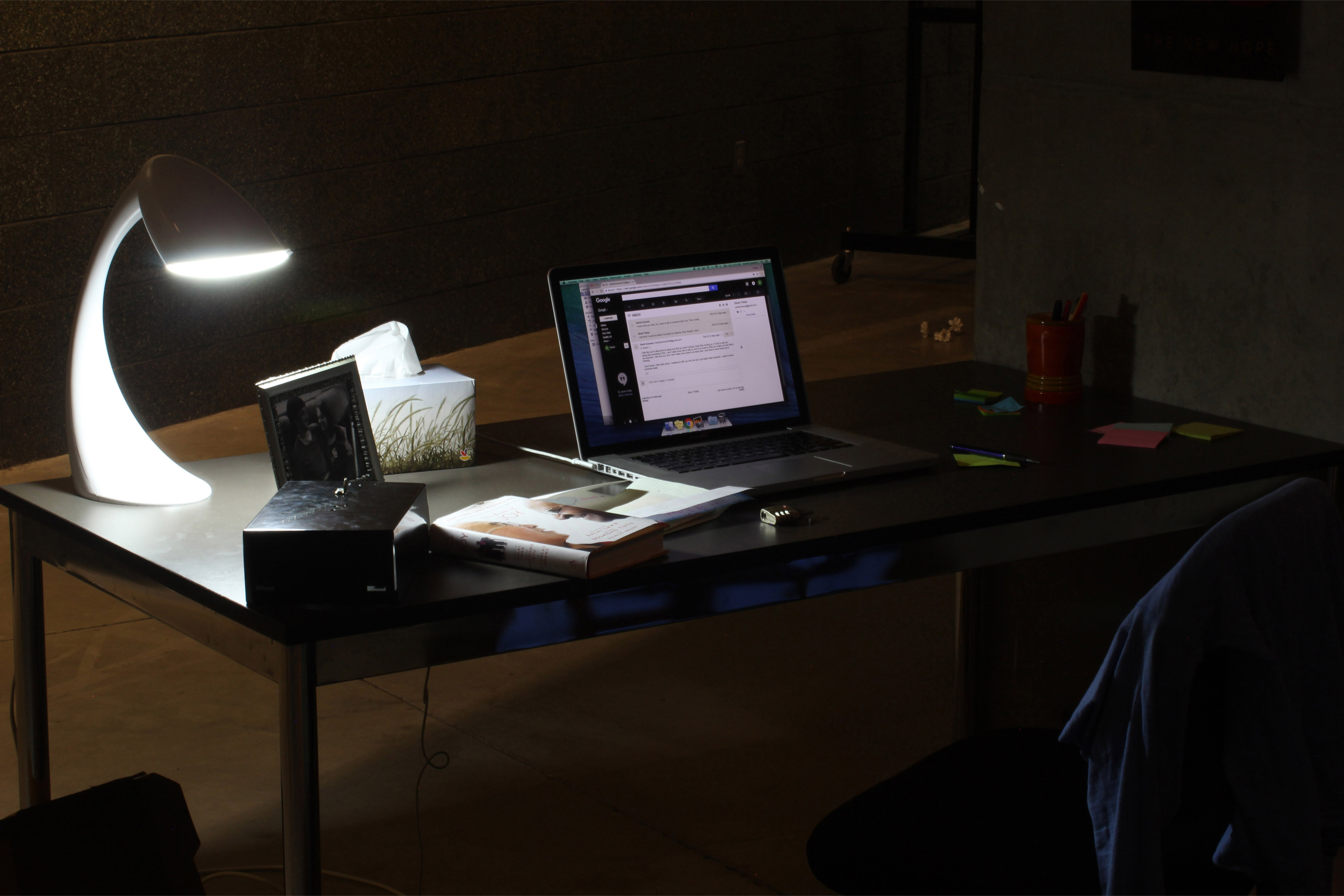 Dark desk dimly-lit by a single lamp. The desk contains a framed photo, tissues, a lockbox, sticky notes, and an open laptop.