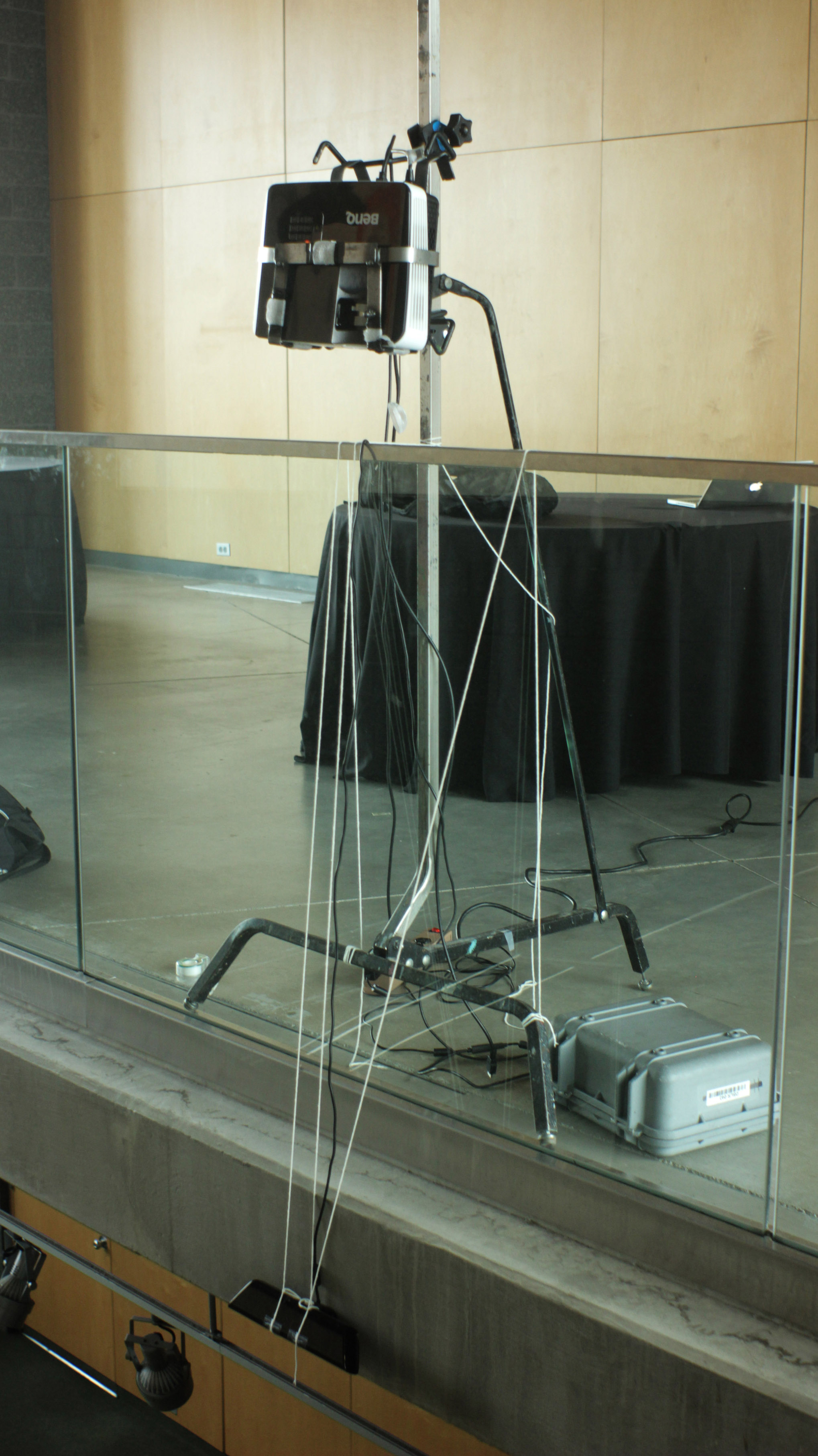 A projector on a stand hangs over a railing, pointing downwards. Strings are attached to the rail, from which a kinect sensor hangs below the projector